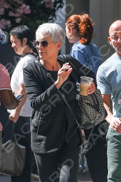 Woman with short gray hair wearing sunglasses and a black outfit, holding a drink while walking in a busy street.