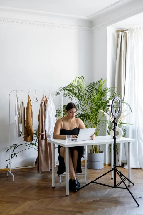 A woman sitting at a desk with a laptop in a stylish, well-lit room. In the background, there is a clothing rack with various outfits and a large plant, along with a ring light set up.