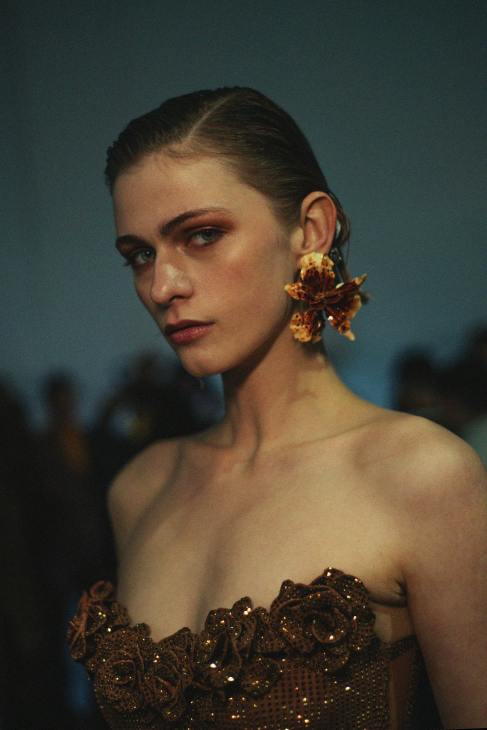A model with a floral earring poses in an elegant, strapless dress featuring intricate floral embellishments.