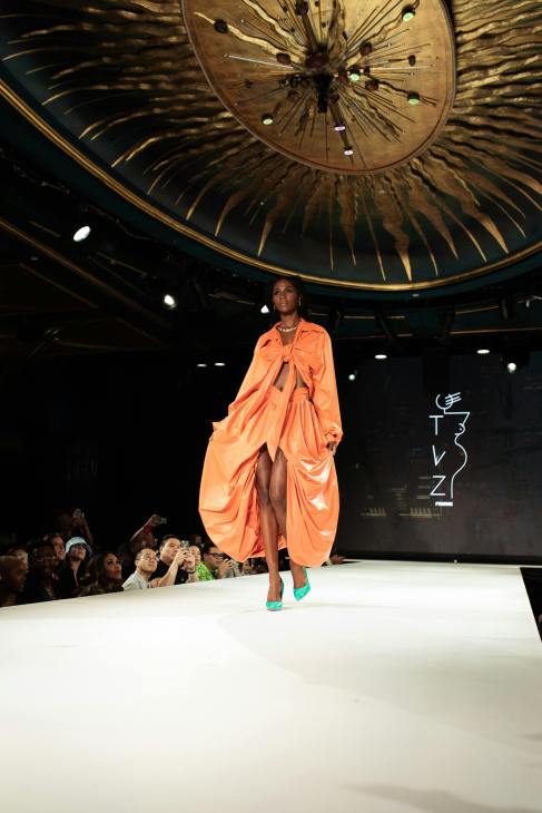 A model walking down the runway in an orange dress with a striking design, showcasing creativity and fashion at a fashion event.