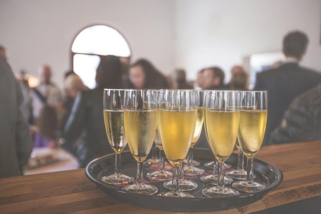 A tray of champagne flutes filled with bubbly drinks, set against a blurred background of an event gathering.