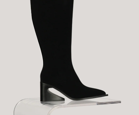 Close-up of a stylish black tall boot displayed on a clear pedestal, featuring a unique geometric heel.