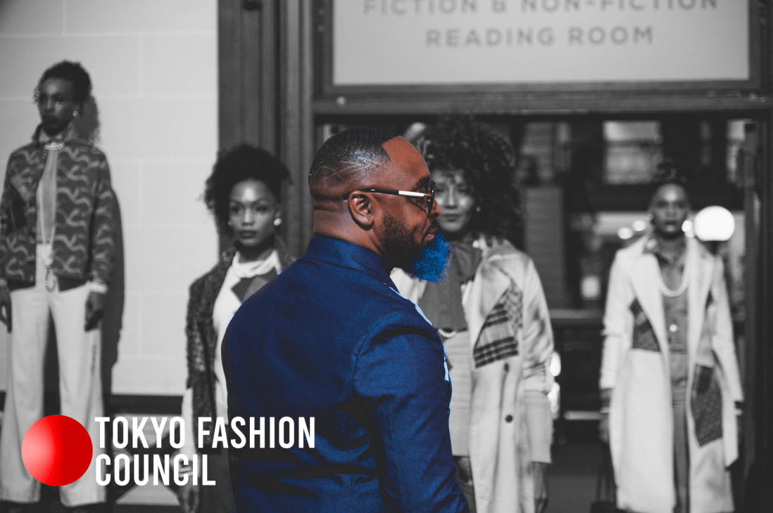 A man with a blue beard and glasses stands in profile, surrounded by models in fashionable attire in the background, with a sign in the upper area indicating a reading room.